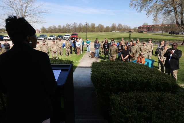 A woman speaks at a podium as several people stand and watch her.