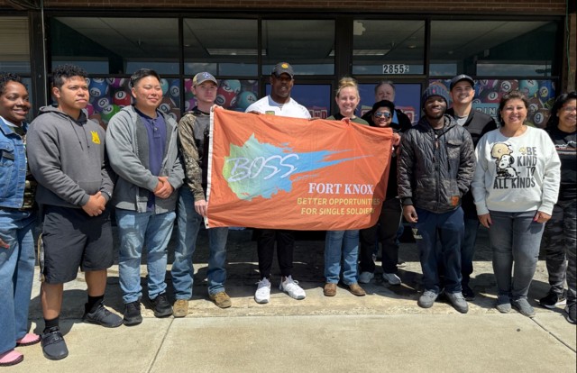 Several people stand beside each other and smile at the camera with three of them holding a brown banner that depicts the shape of Kentucky and reads, &#34;BOSS Fort Knox.&#34;