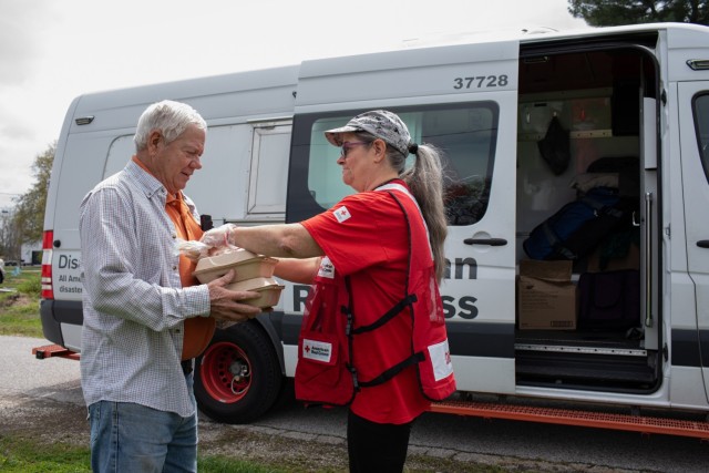 A woman gives a man two Styrofoam containers of food with a Red Cross van parked behind them.
