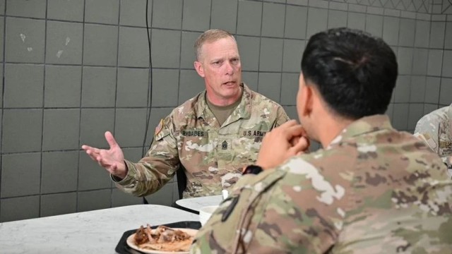 A man sitting in a chair seated at a table gestures with his right hand, mouth open, as he looks at a man sitting across from him.