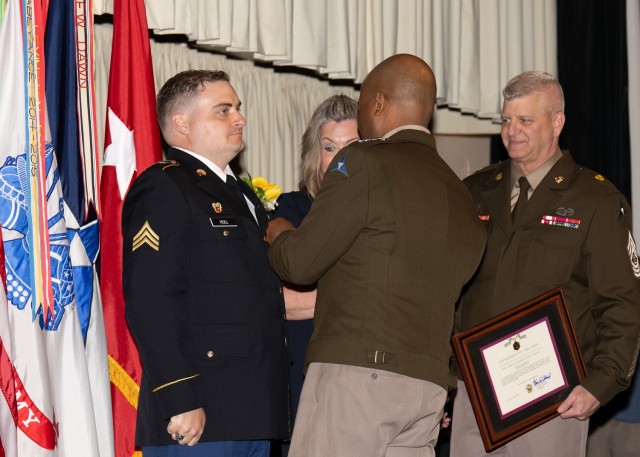 A man pins a medal on another man&#39;s suit, standing in front of him, while two people on the side, a man and a woman, look on.