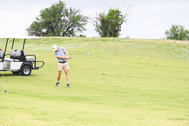 A man looks down at the ground, a golfing iron in his hands mid-swing, and a golf ball in the air several feet in front of him.