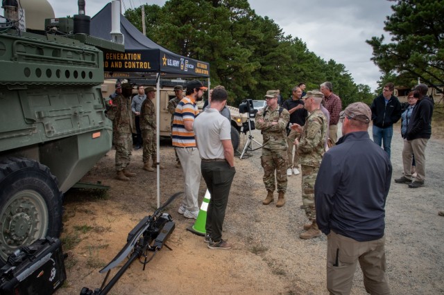 Soldiers and civilians come together for an information briefing in front of a modified Stryker and a drone against Uas.