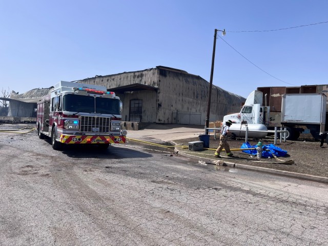A fire truck sits on the street in front of a large, fire-damaged metal building.