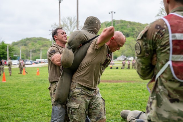 A man standing grips a piece of rope wrapped around a sandbag dummy propped against his back as another man standing behind the dummy pushes it up the man's back.