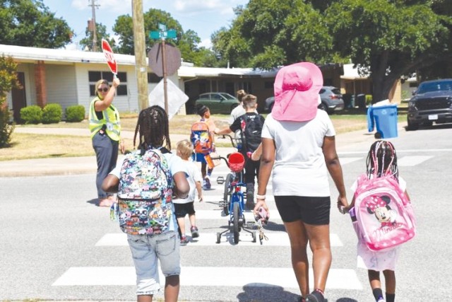 Children and adults walk across a crosswalk, the children carrying backpacks on their backs.