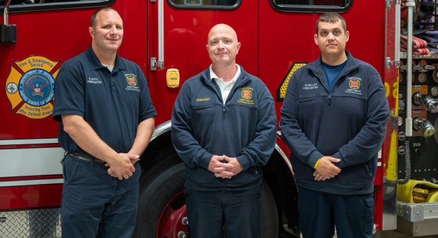 Three people wearing Tobyhanna Army Depot Fire & Emergency Services apparel stand in front of a firetruck and pose for a photo.