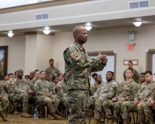 A man stands with his hands out in front of him and mouth open. In front and to the left of him, people sit in chairs in rows and look on.