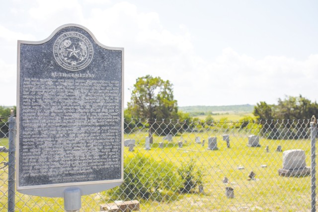 A Stone and metal marker stands in front of a grassy field with headstones in rows.