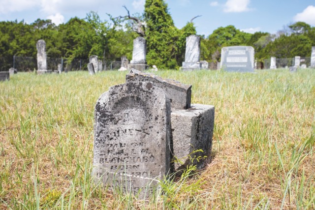 A piece of a headstone sits against its base in a grassy field. Behind, other headstones sit in rows.