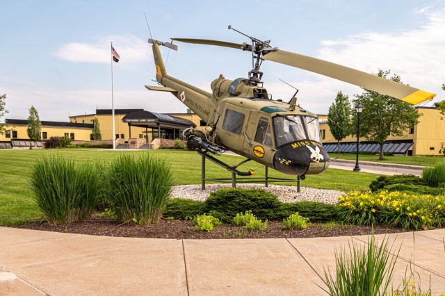 A helicopter is displayed on the front lawn of a building with a large flagpole flying an American and POW/MIA flag in the background.