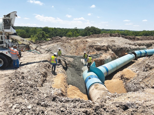 Four people standing surround a metal, open chute pouring wet concrete on the ground. A man stands by the end of a vehicle to which the tube is attached.