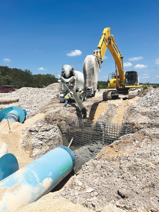 Two men push on opposite sides of an open metal chute pouring concrete into a large hold in the ground. Beside them, an excavator sits.
