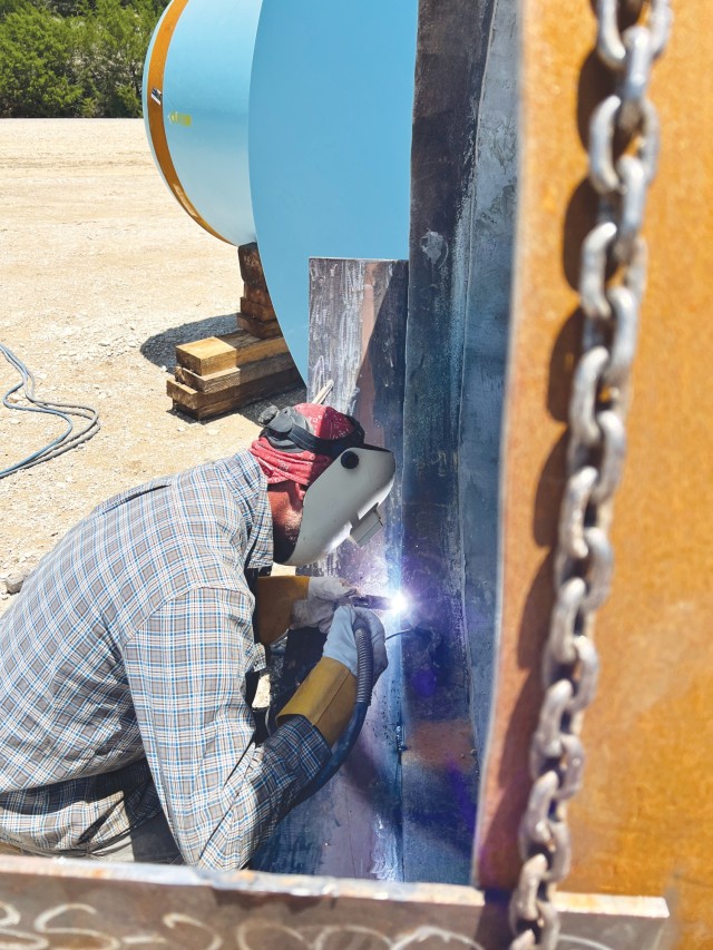 A man crouches over the edge of a circular metal rim wearing a metal face cover and thick gloves while holding welding equipment, sparks emitting where the tool is touching the metal.