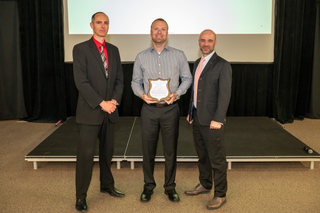 Three people pose for a photo during an awards ceremony. The person at center holds a plaque.