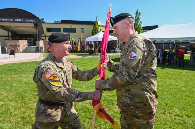 Two people wearing military uniforms participate in an outdoor change of command ceremony. One passes a flag to another.