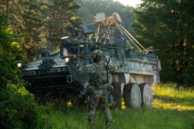 A soldier ground guides a Stryker fitted with CUAS equipment in the woods of Hohenfels, Germany, for a training exercise.