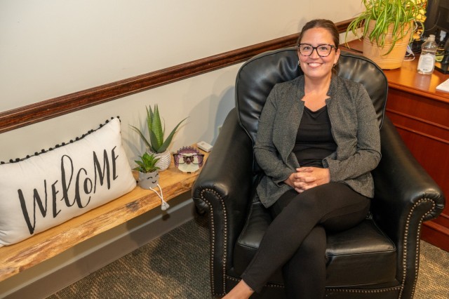 A person poses for a photo in a chair near a shelf with plants and a welcome pillow on it.