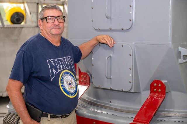 A person wearing a Navy shirt poses for a photo next to a military asset.