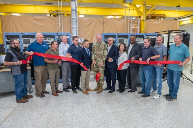 13 people hold a ribbon in front of equipment. The people at center cut the ribbon.