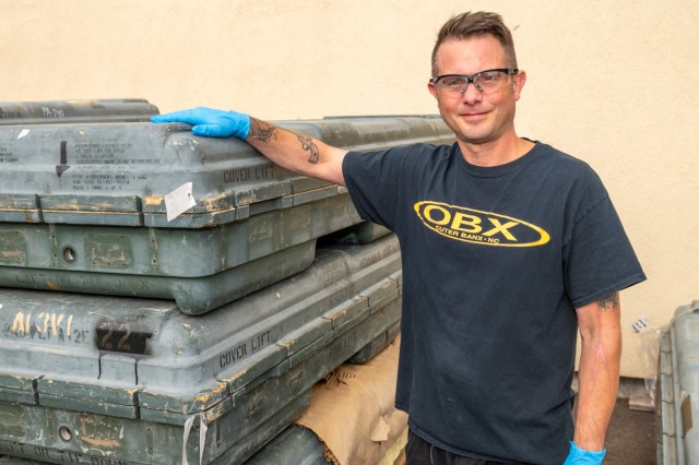 A person poses for a photo in a production floor area. The person is wearing gloves and rests their hands on a case