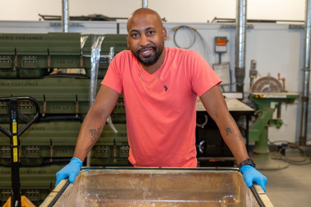 A person poses for a photo in a production floor area. The person is wearing gloves and holds a case