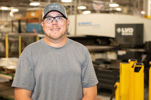 A person poses in front of industrial machinery for a photo. They are wearing a hat and safety glasses.