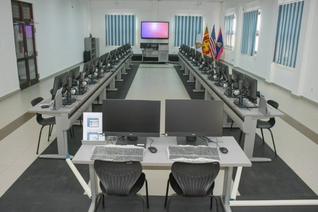 A computer lab - several computer monitors, keyboards, and mice sit on tables. A smart board and flags are seen in the background.