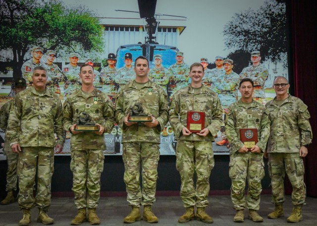 Six soldiers are standing in a line, shoulder to shoulder. four of them are holding awards - two are trophies, two are plaques. Those four are framed on either side by a command leadership