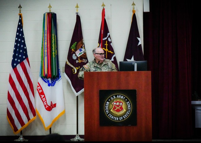 A two-star general officer speaks from behind a podium with five flags behind him.