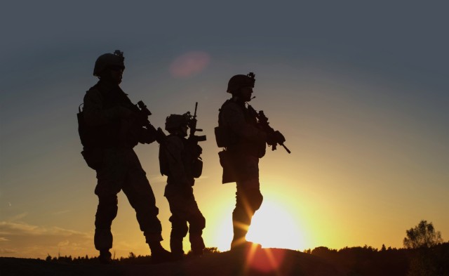 Squad of Three Fully Equipped and Armed Soldiers Standing on Hill in Desert Environment in Sunset Light.