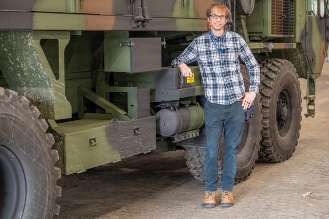 A person poses for a photo resting their arm on a military vehicle.
